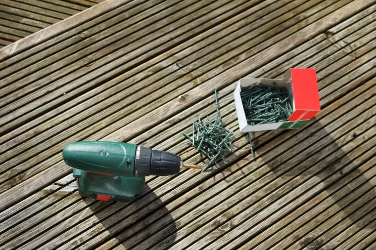 Perceuse-visseuse verte posée sur une terrasse en bois, à côté d'un tas de vis et de leur boîte