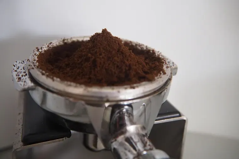 Porte-filtre rempli de café moulu en monticule, posé sur un support, devant un mur clair